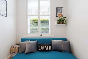 Ein Zimmer mit einem blauen Sofa, einem Fenster mit Jalousien, einem kleinen Regal mit Pflanzen und einem gerahmten Bild an der Wand.