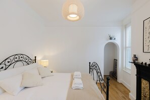 Un dormitorio con cama, lámparas de noche, una lámpara colgante, chimenea y una planta pequeña en un estante.
