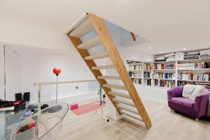 Ein Zimmer mit einer Wendeltreppe aus Holz, einem Glastisch, einem violetten Sofa und einem Bücherregal voller Bücher.