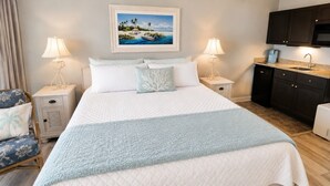 A bedroom with a bed, two bedside tables with lamps, a chair, a painting on the wall, and a kitchenette.