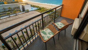 A balcony with two wicker chairs, a view of the beach, and a parking lot.