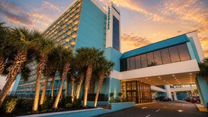 A hotel with a modern design, featuring a prominent tower and a large entrance with a parking area.