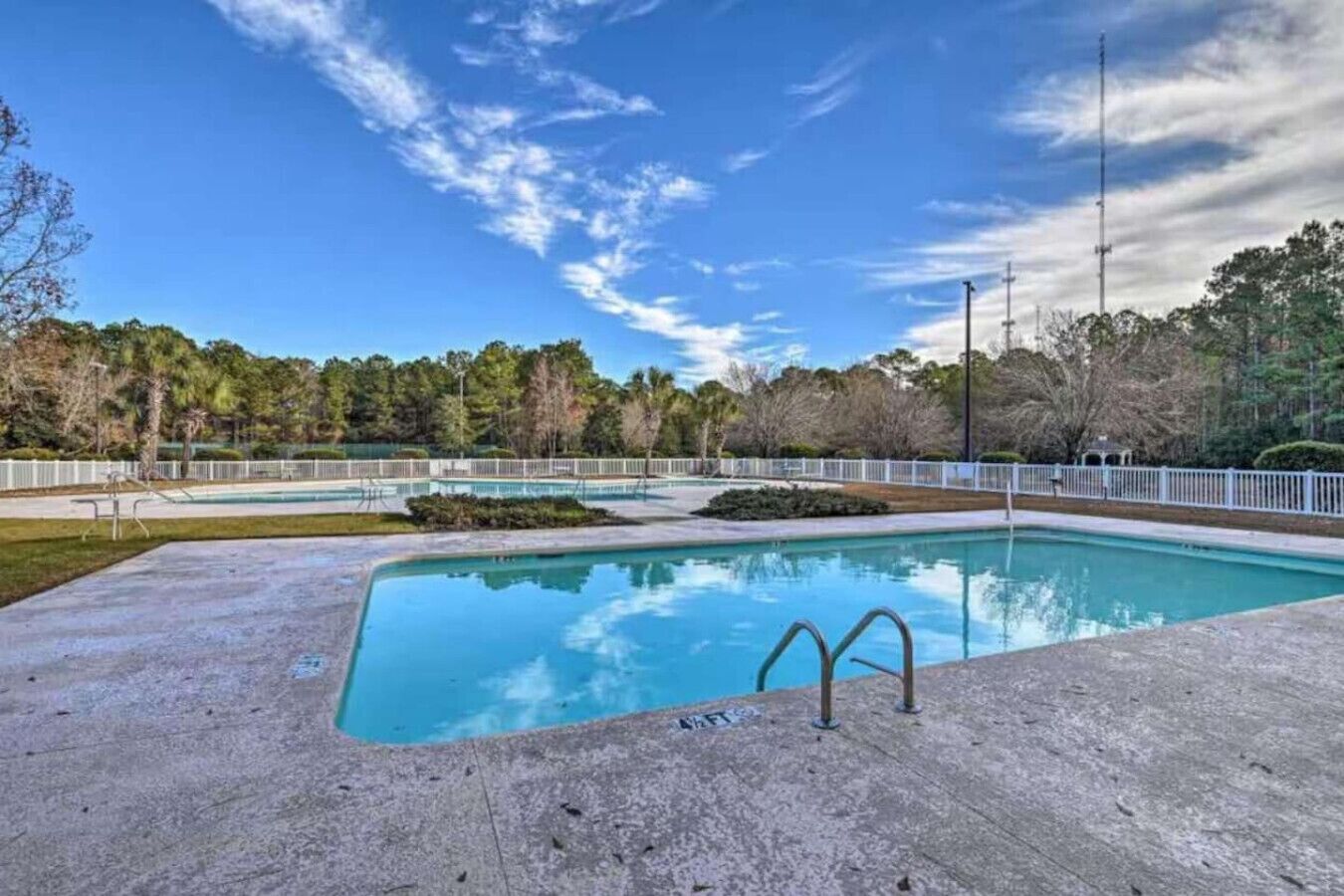 A rectangular swimming pool with a concrete deck, surrounded by a white fence, and trees in the background.