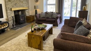 Ein gemütliches Wohnzimmer mit Kamin aus Stein, einer Sofagarnitur, einem Holztisch und Blick durch ein großes Fenster.