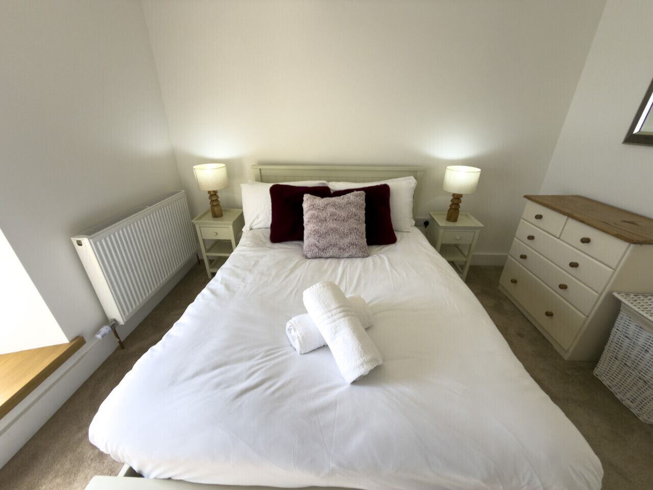 A neatly made bed with white linens, a red pillow, and two rolled towels. There are bedside tables with lamps, a radiator, and a wooden dresser with drawers.