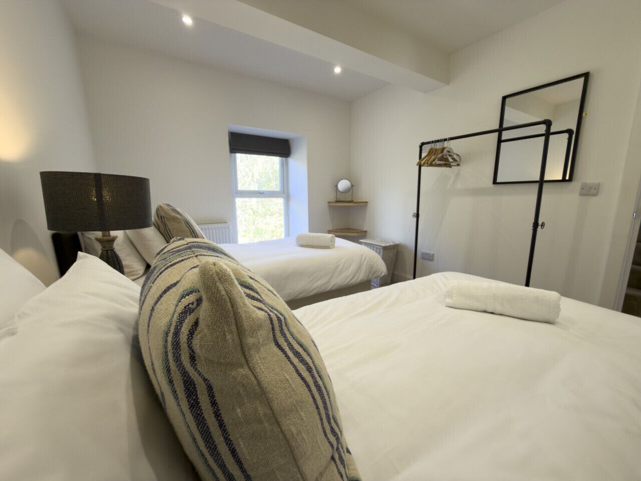 A hotel room with two beds, a bedside table, a lamp, a mirror, and a window.