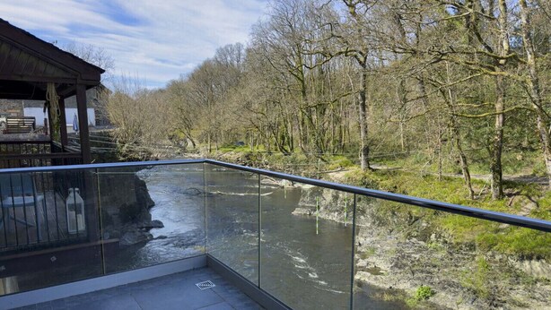 Ein Balkon am Flussufer mit Glasgeländer, ein Steinhaus im Hintergrund und Blick auf den Fluss und Bäume.