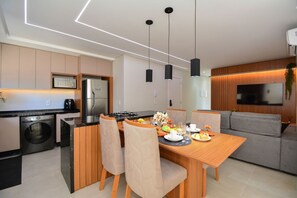 A modern kitchen with a built-in washer, a dining area with a wooden table and chairs, and a grey sofa. - Aconchego Frente Mar em Palmas - GCR010 (Governador Celso Ramos)