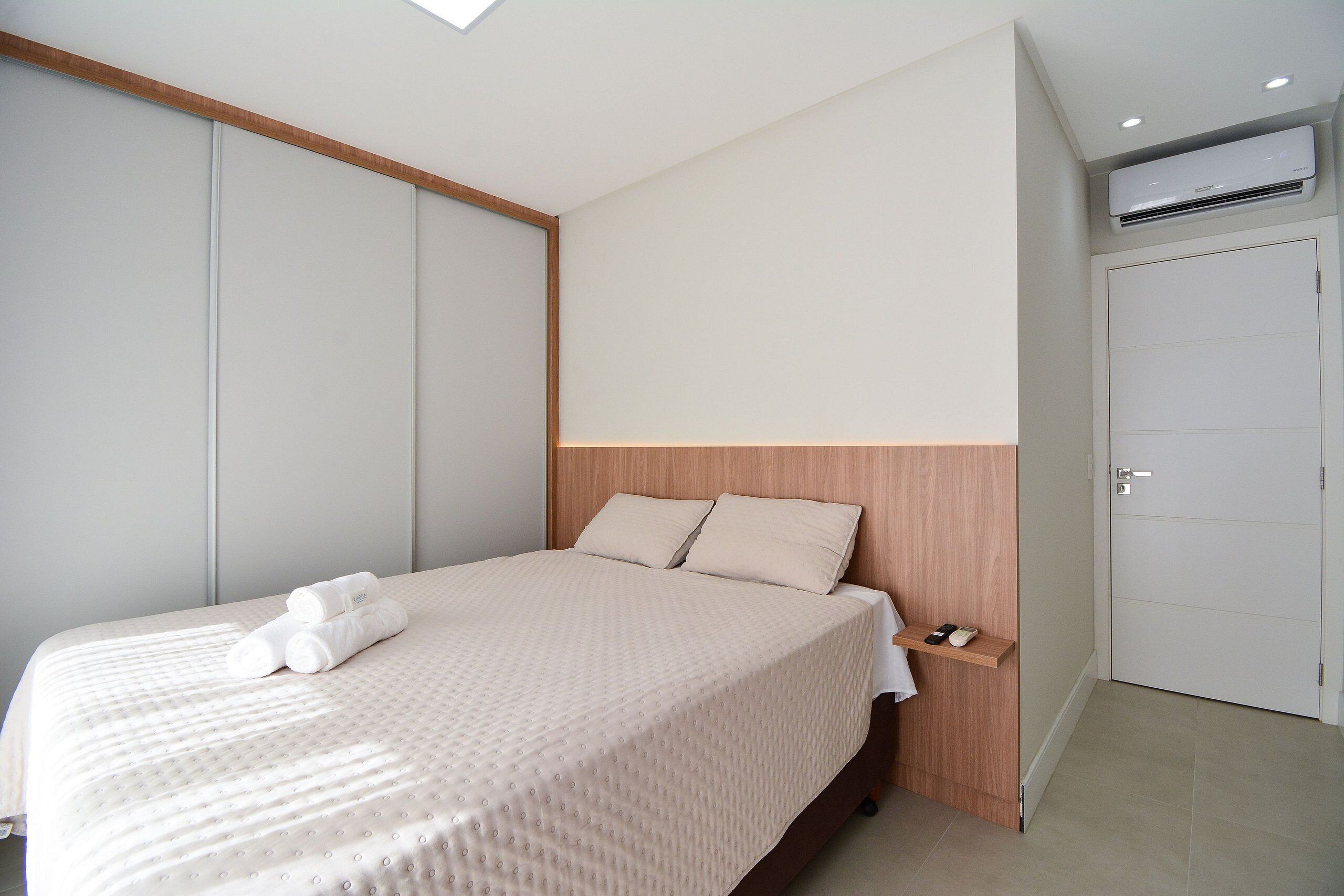 A neatly made bed with a wooden headboard, a bedside table with a remote control, and a wall-mounted air conditioner.