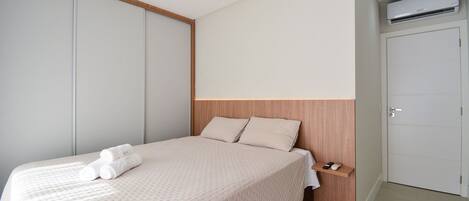 A neatly made bed with a wooden headboard, a bedside table with a remote control, and a wall-mounted air conditioner.