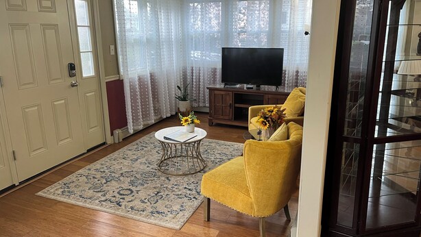 A living room with a yellow armchair, a round coffee table, a TV, and a large window with curtains. - Limbe Guest House in Latham NY (Latham)