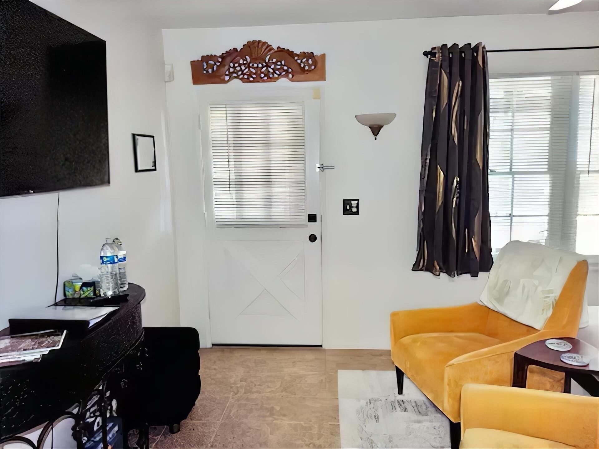 Ein Zimmer mit Schreibtisch, einem Sessel, einer Couch, einer Tür mit dekorativer Verkleidung, einer Wandleuchte und einem Fenster mit Jalousien.