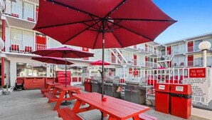 Zona junto a la piscina con mesas de picnic rojas y sombrillas, una parrilla, contenedores de basura y un cartel con el horario de la piscina.