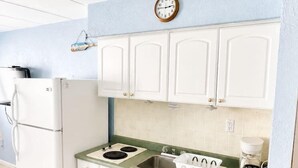 Cocina compacta con armarios blancos, estufa, fregadero, microondas y un reloj en la pared.