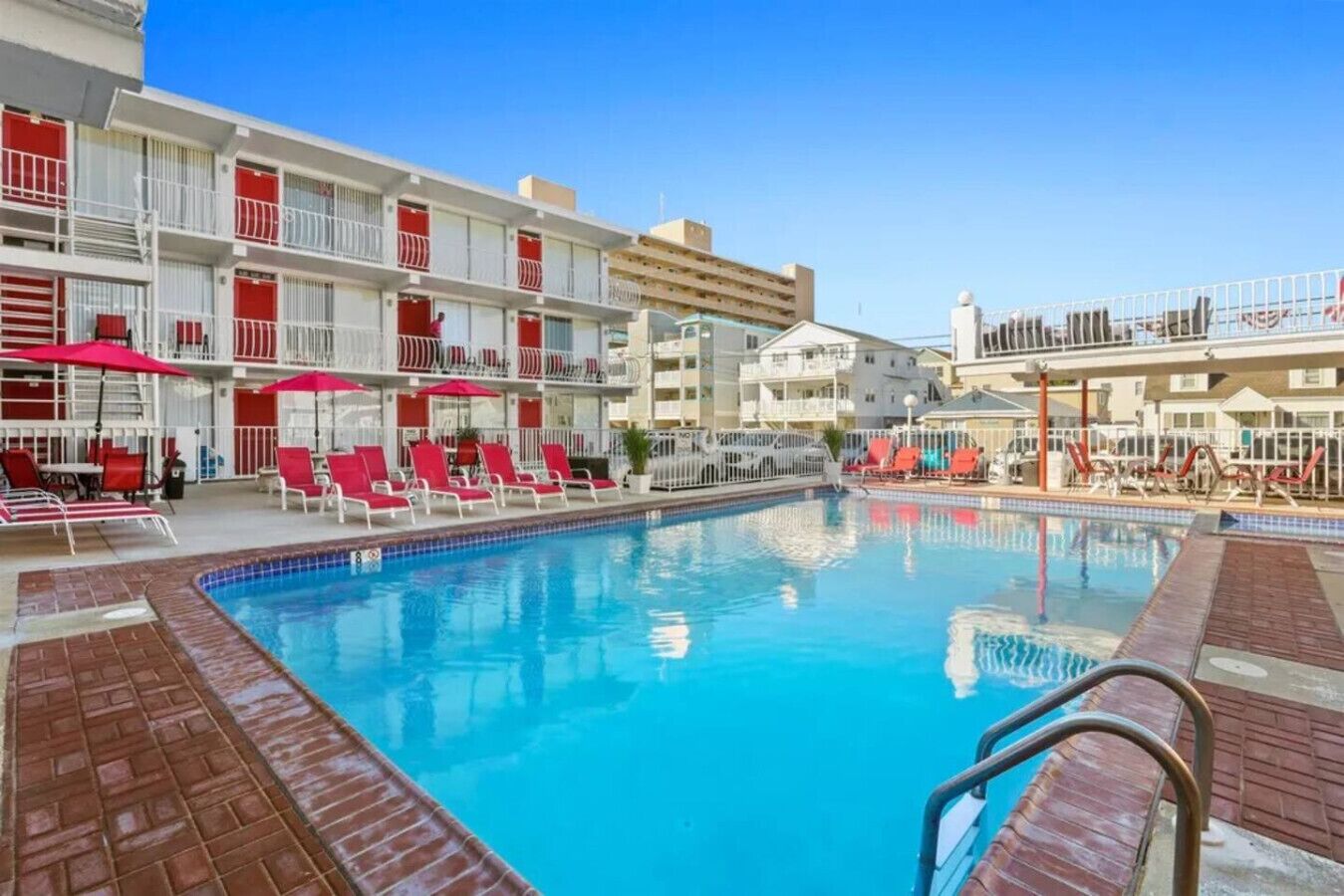 A swimming pool with red lounge chairs and umbrellas, surrounded by multi-story buildings with balconies.