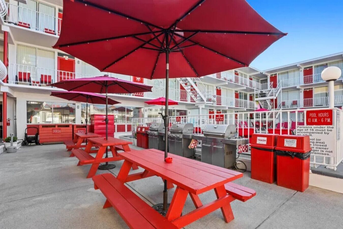 A poolside area with red picnic tables and umbrellas, a barbecue grill, and trash bins.
