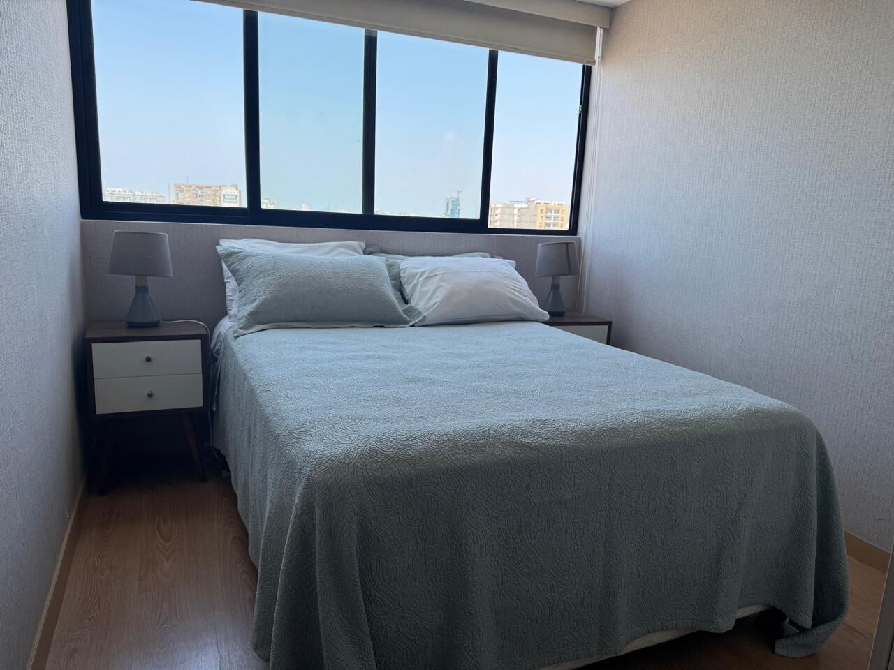 Un lit bien fait, recouvert d’une couvre-lit verte, deux tables de chevet avec des lampes et une grande fenêtre donnant sur la ville.