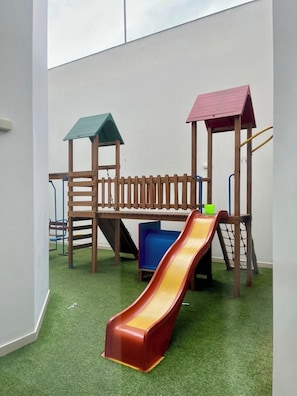Une structure de jeu en bois, dotée d’un toboggan et d’éléments d’escalade, installée sur une surface de gazon synthétique.