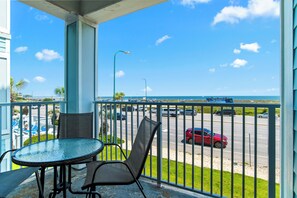Ein Balkon mit Tisch und Stühlen, der auf eine Straße mit Autos und den Blick aufs Meer zeigt.
