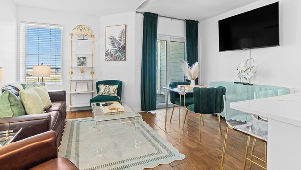 Una sala moderna con un sofá de cuero, una mesa de centro de vidrio, un sillón color turquesa, un televisor montado en la pared y un ventilador de techo.