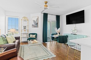 Una sala moderna con un sofá de cuero, una mesa de centro de vidrio, un sillón color turquesa, un televisor montado en la pared y un ventilador de techo.