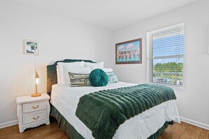 Ein Schlafzimmer mit Bett, Nachttisch, Lampe, einem Fenster mit Jalousien und einem gerahmten Bild an der Wand.