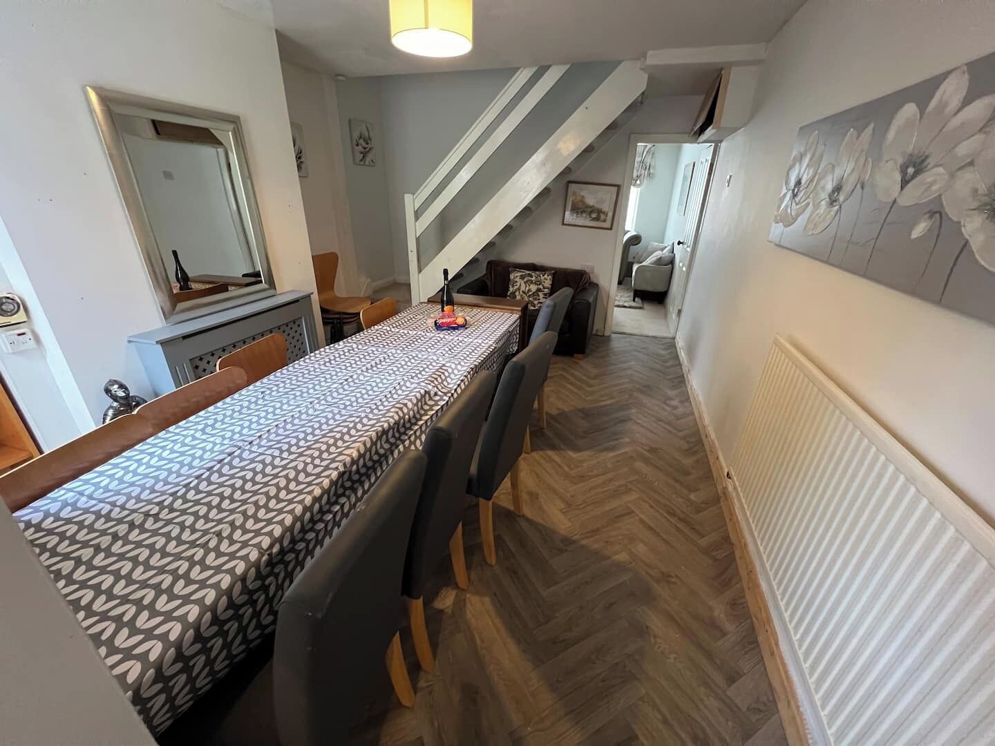 Un comedor con mantel estampado, sillas de madera, un radiador y una escalera que sube a otra habitación.