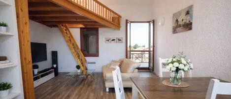 Un amplio salón con escalera de madera, un sofá, una mesa de comedor con sillas y un balcón con vistas.