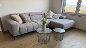 Un salon moderne avec un canapé d’angle gris, deux petites tables rondes en fil de métal et une vue sur un paysage vallonné par la fenêtre.