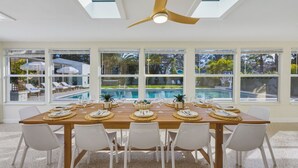 A dining area with a long wooden table set for six, large windows with a view of a swimming pool, and a ceiling fan. - NEW - Sweet St Pete - 5 Minutes to beach - Heated Pool, Hot Tub, Sauna, + more (St. Petersburg)