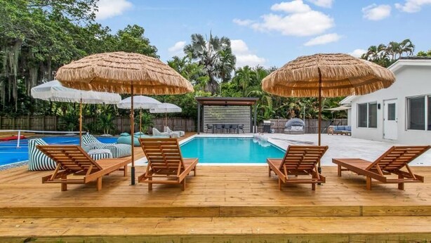 A pool area with lounge chairs and thatched umbrellas, a covered seating area, and a house in the background. - Boho Villa by Glam Homes (Miami)