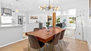 A modern kitchen with a central island, a dining area with a wooden table and chairs, and a living space with a sofa and a large window. - Boho Villa by Glam Homes (Miami)