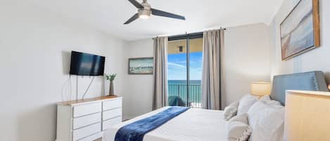 Et hotelværelse med seng, ventilator, fjernsyn, et vindue med gardiner og et billede på væggen.