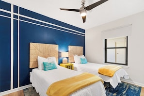 Quarto de hotel com duas camas, um ventilador de teto, uma janela com persianas e uma parede com padrão geométrico.