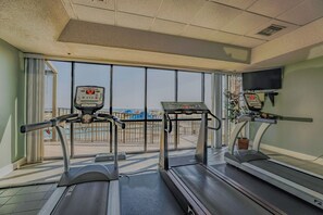 Ein Fitnessraum mit Laufbändern, einem Ergometer und einem großen Fenster mit Blick auf den Strand.