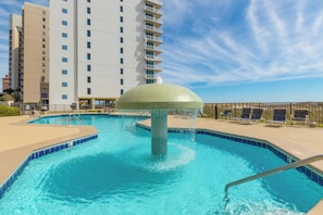 Piscina com uma fonte de água, rodeada por espreguiçadeiras e um edifício alto ao fundo.