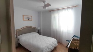 Ein Schlafzimmer mit einem Bett, einem Deckenventilator, einem Nachttisch, einer Lampe und einem Fenster mit Vorhängen.