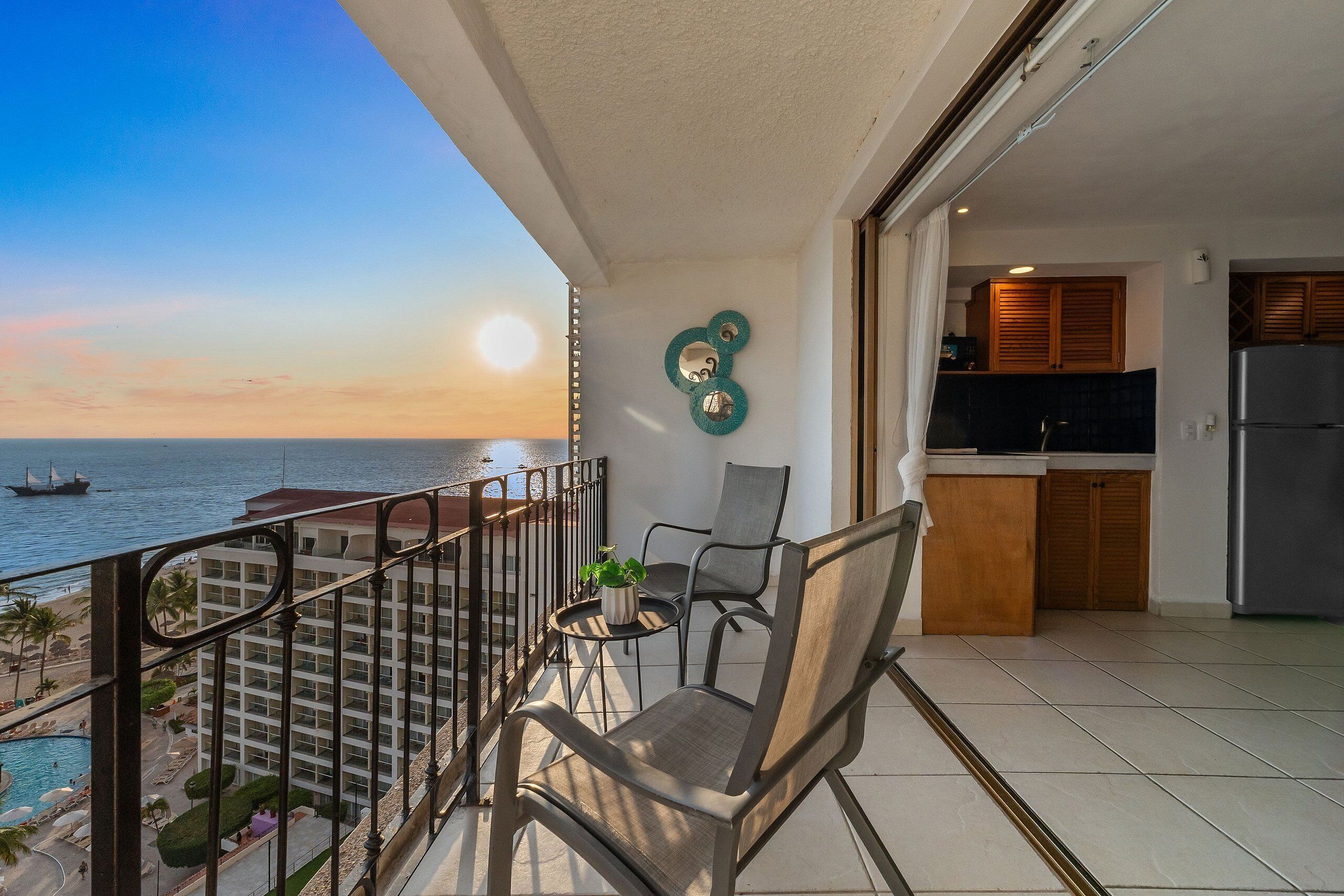 Un balcón con vista al mar, una silla, una mesita con una planta y una zona de cocina.