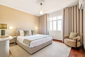 Ein Hotelzimmer mit Bett, Sessel, Nachttisch, Lampe und einem Fenster mit Vorhängen.