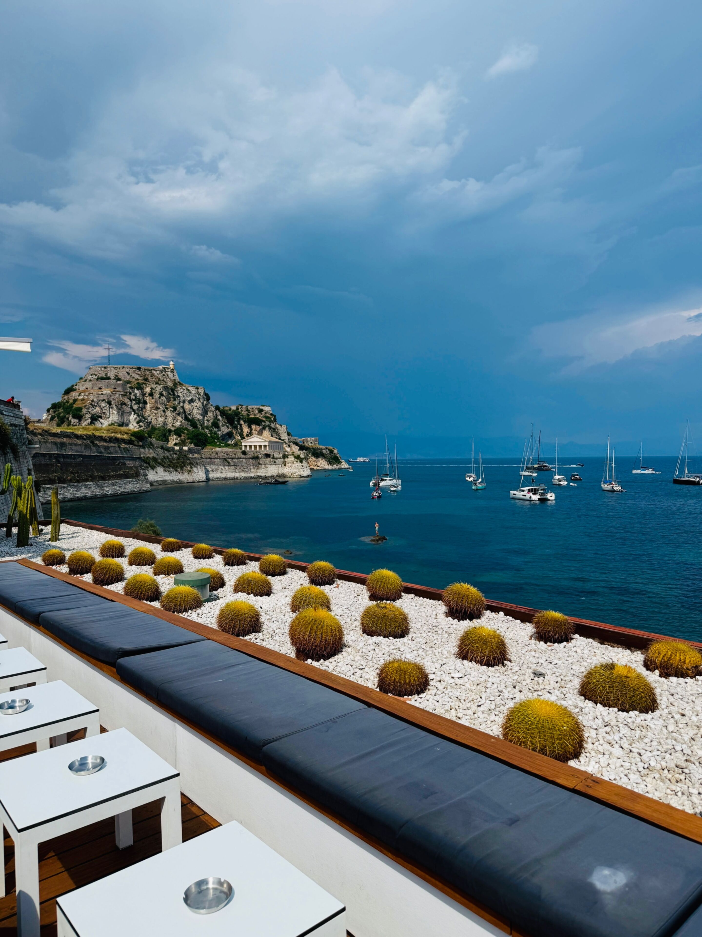 Une terrasse en bord de mer avec des tables et des coussins blancs, une falaise rocheuse sur laquelle se trouve un bâtiment, et des voiliers sur l’eau.