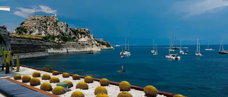 A seaside terrace with white tables and cushions, a rocky cliff with a building, and sailboats on the water.