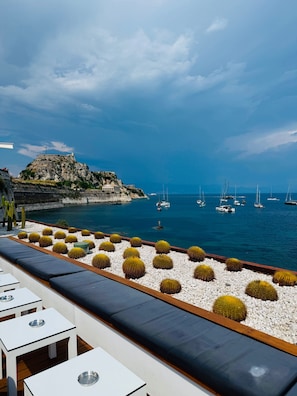 A seaside terrace with white tables and cushions, a rocky cliff with a building, and sailboats on the water. - Art House Seaview & Jacuzzi Spartylas Sleeps 4 (Corfu)