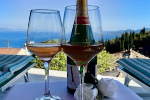 Two wine glasses on a table with a bottle of wine, a white napkin, and a view of the sea and hills in the background. - Art House Seaview & Jacuzzi Spartylas Sleeps 4 (Corfu)