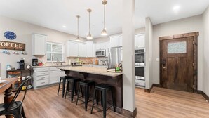 Una cucina moderna con mobili bianchi, un'isola centrale con sgabelli, elettrodomestici in acciaio inossidabile e illuminazione a sospensione.