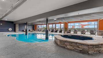 An indoor swimming pool with lounge chairs and a hot tub, large windows, and a stone wall.