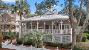 A white colonial-style house with a porch, surrounded by a white picket fence and palm trees.