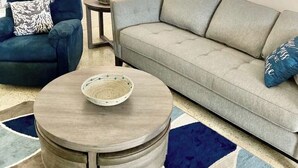 Uma sala de estar com um sofá cinzento, uma poltrona azul, uma mesa de centro redonda e um tapete estampado.