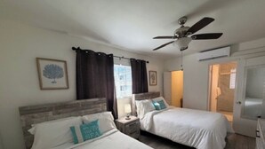 Quarto de hotel com duas camas, um ventilador de teto, uma janela com cortinas e uma porta de acesso a um banheiro.