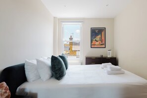 A hotel room with a bed, pillows, a nightstand, a painting, and a view through the window. - Gorgeous 1BDR Flat in the Heart of Clapham (Greater London)