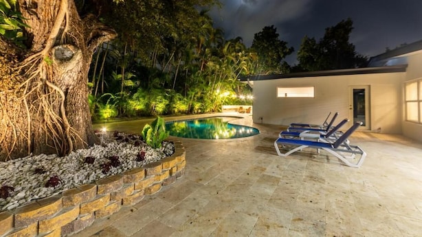 A swimming pool with lounge chairs, a tree, and a building with a lighted exterior. - Villa Azur by Glam Homes (North Miami Beach)